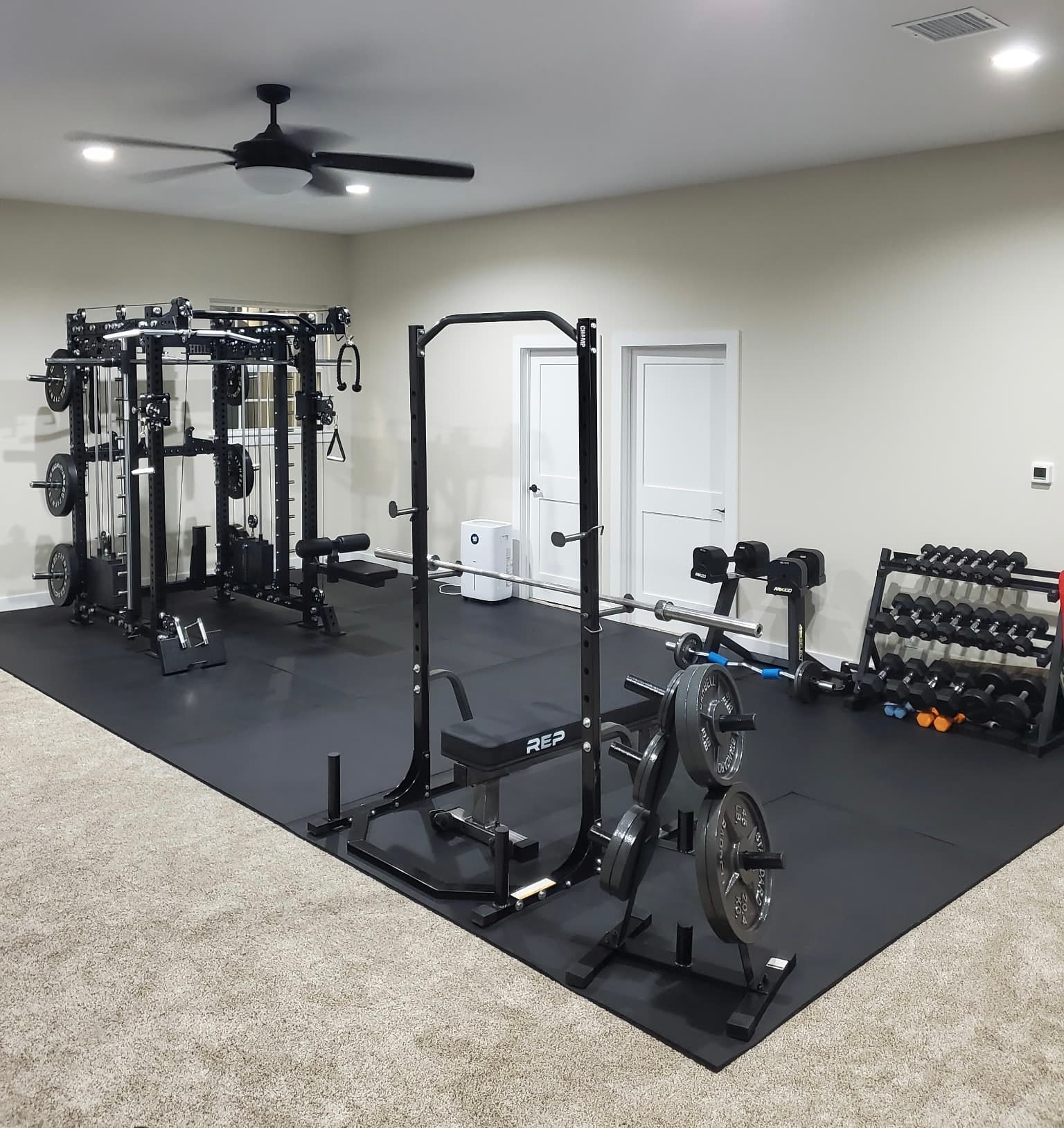 Well-equipped home gym with a power rack, cable machine, and dumbbells on black mats.
