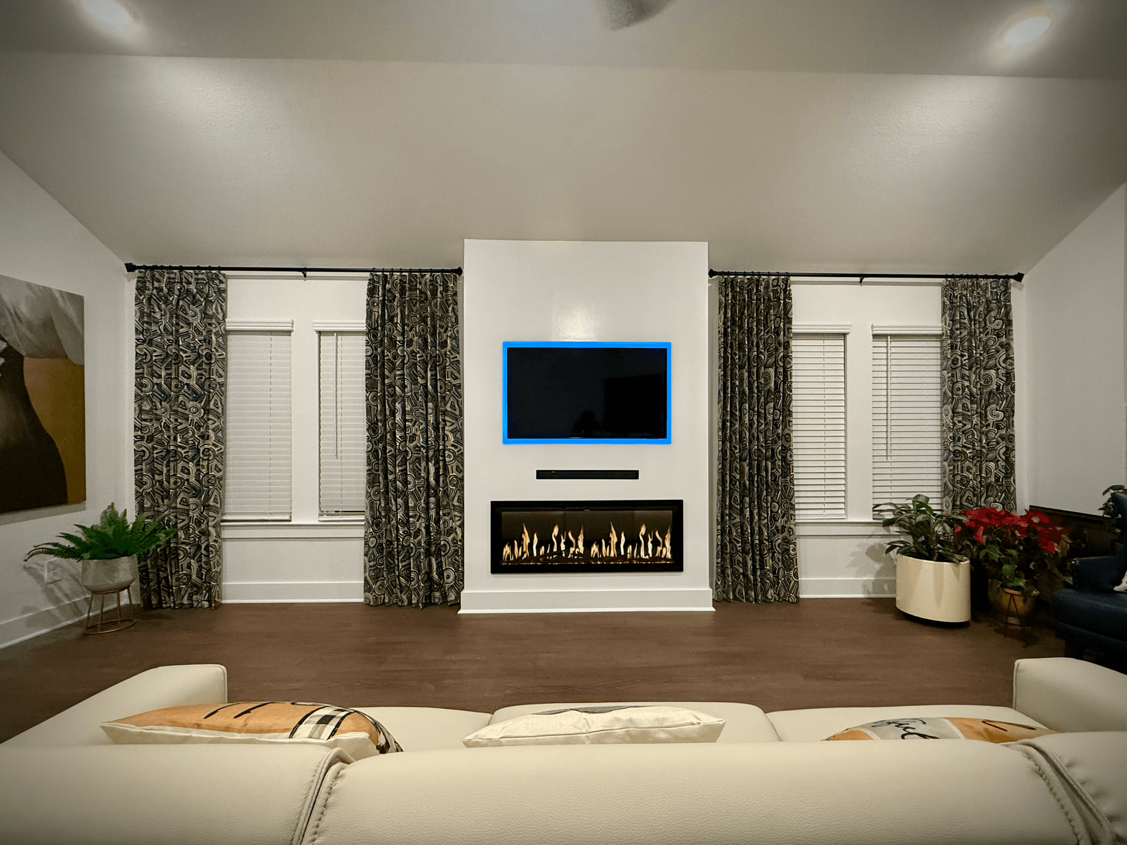 Modern living room with a central fireplace, blue-backlit TV, patterned curtains, and white sofa.