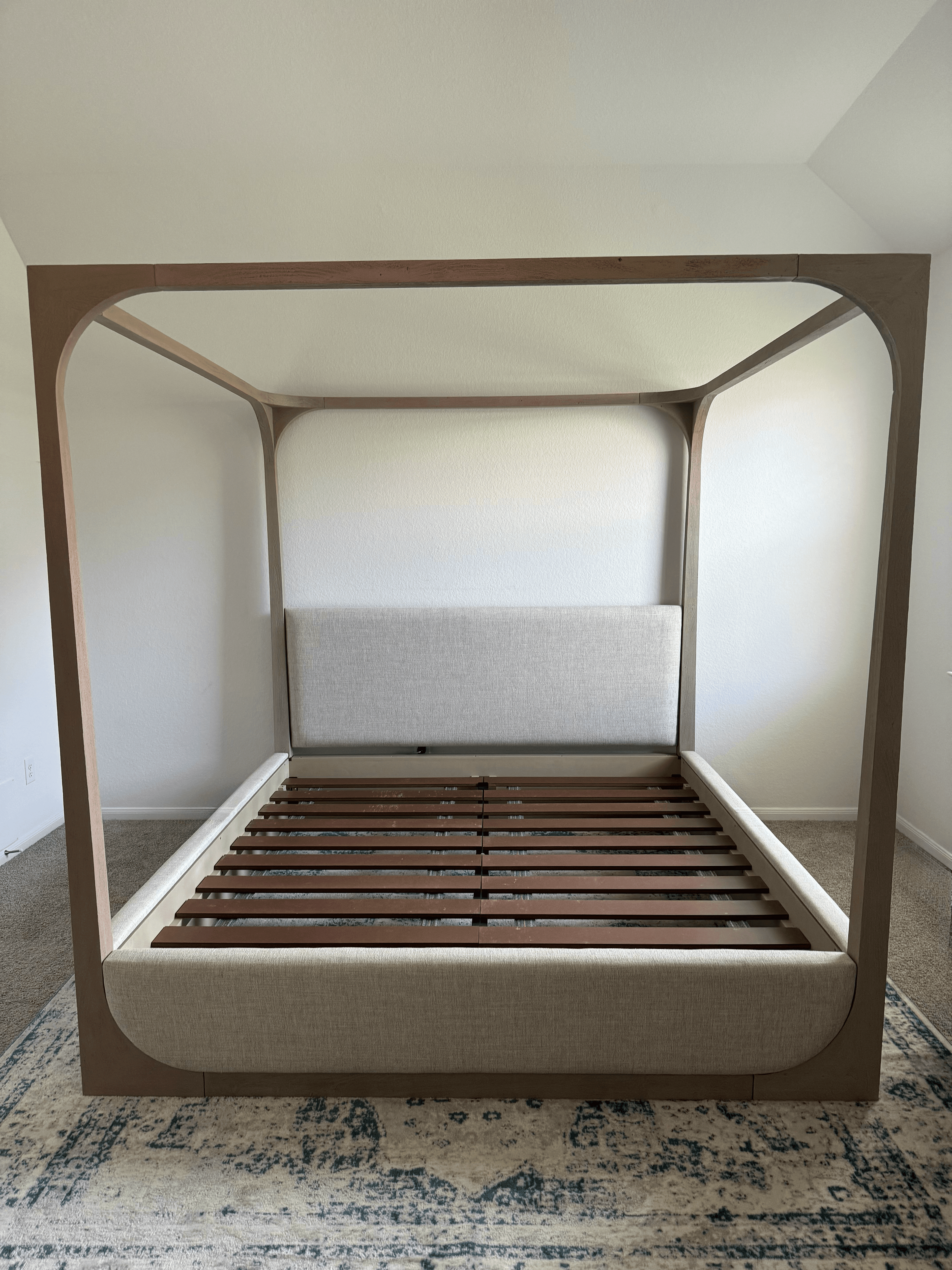 Modern light wood canopy bed frame with upholstered headboard and base on a patterned rug.