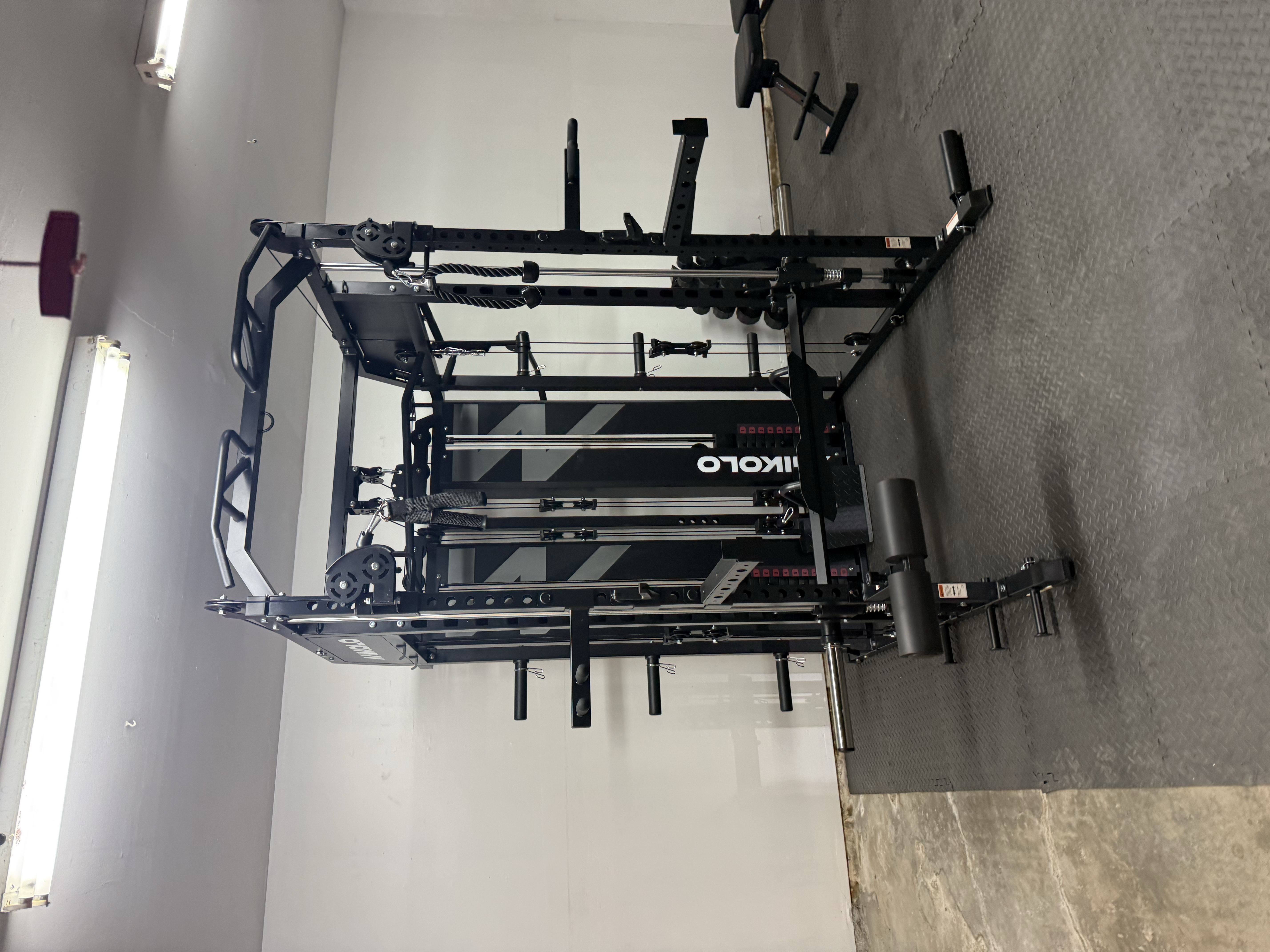 Black Mikolo power rack with Smith machine and cable pulleys on rubber gym flooring.