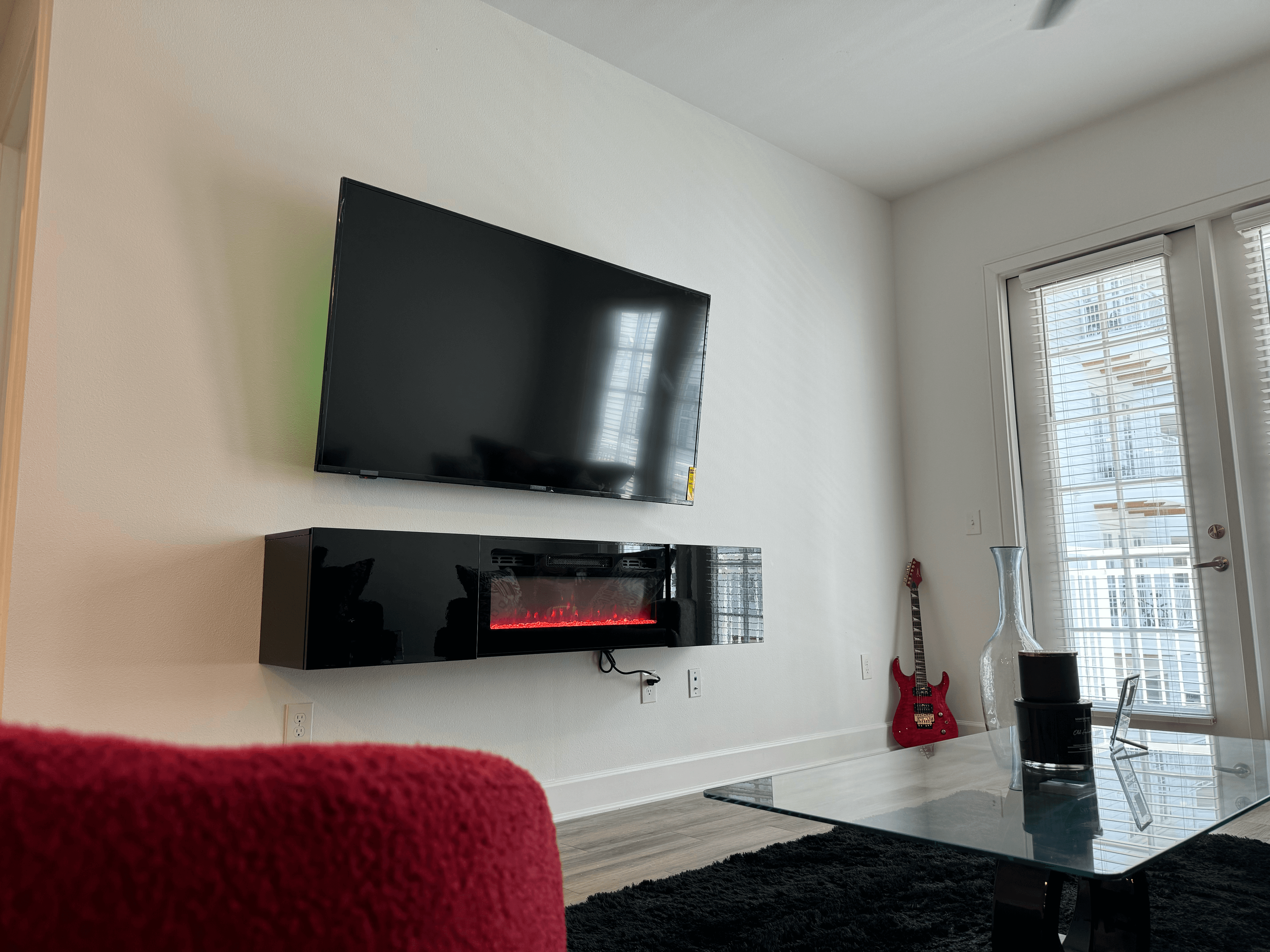 Modern living room featuring a wall-mounted TV, electric fireplace, red guitar, and glass table.